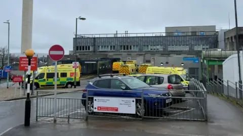 Ambulances and a couple of cars parked outside Derriford Hospital in Plymouth. It is a cloudy and wet day. The hospital building is grey.
