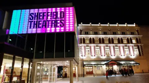 Sheffield Theatres The modern Crucible Theatre has large panels on a balcony lit up in rainbow colours. In the background is the historic Lyceum Theatre lit up by white lights