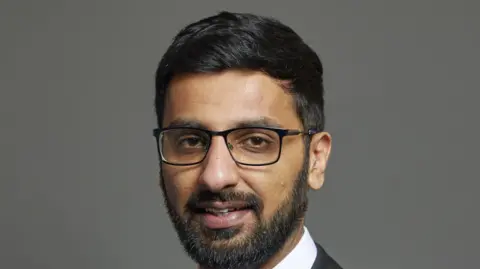 Parliament UK Official portrait of Adnan Hussain taken against a grey backdrop. He has black hair and a beard and is wearing glasses.