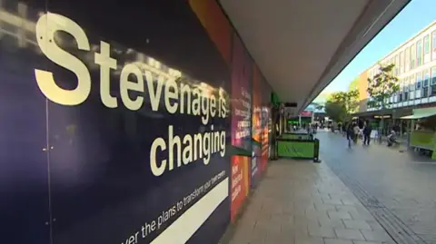 BBC Shopping street in Stevenage. A banner on the wall to the left indicates that "Stevenage is changing". There is a cafe further down the street with green barriers either side of tables and chairs . There is a green food stall to the right in front of three-storey modern shop buildings. Some shoppers can be seen walking on the block-paved pavement.