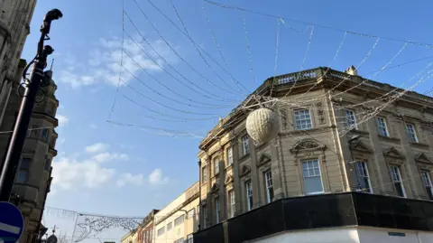 Above the cross where four pedestrianised high streets meet, a large silver ball sits in the middle of a web made of lights which has been strung between buildings. Silver wire banners of Christmas decorations can be seen hanging down one of the streets. It is a sunny day in the spring with minimal cloud in the blue sky and historic architecture towering over the streets.