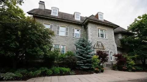 An image showing the outside of 24 Sussex Drive. It is a medium-sized cottage with a grey brick facade, a large birch tree next to the front door, which is dark green and is surrounded by an ornate arch with three short steps in front of it. 