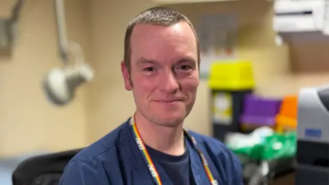 Un sonriente Dr. Merriel con matorrales azules del NHS y tiene una insignia del NHS alrededor de su cuello en colores del arco iris. Está sentado en su cirugía de GP y hay equipos médicos en el fondo. 