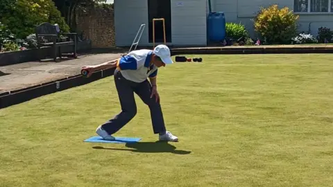 A person bends down to throw a bowl on a short green lawn in bright sunshine.