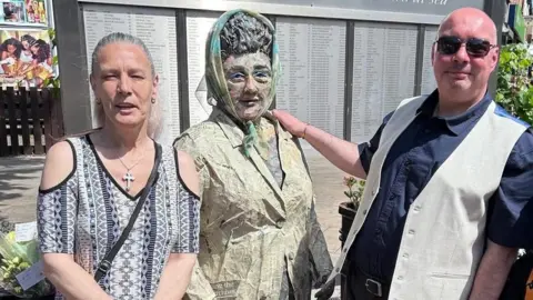 Councillor Tracey Henry, chair of the Headscarf Revolutionary statue management committee, and Ian Cuthbert with a life-sized paper sculpture of Lillian Bilocca. Henry is wearing a purple and white patterned top with cut-out shoulders, a silver cross necklace and a black bag across her body. She has long, grey hair tied back in a ponytail. Cuthbert is bald and is wearing black sunglasses, a navy shirt with the sleeves rolled up and a beige waistcoat undone. 