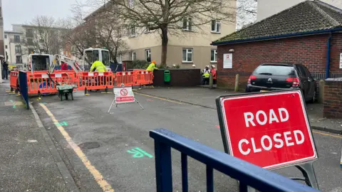 BBC A red ROAD CLOSED sign in front of a fenced off part of a road surrounded by residential buildings with people in high vis gear working.
