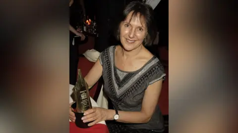 Getty Images A woman seated at a table holding an award shaped like the nib of an ink pen.  She has dark hair and brown eyes and is smiling at the camera.  She is wearing a grey top embellished with silver and black shiny beads
