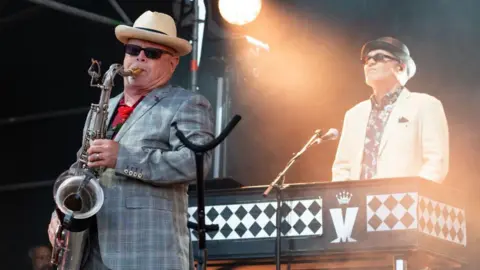 Madness performing at Uptown Festival Blackheath 2024 at Blackheath Common in London. Lee Thompson is on the saxophone and Mike Barson is on the keyboard. Both are wearing hats and sunglasses.