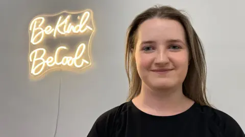A young woman with long dark brown hair, wearing a black t-shirt and smiling at the camera. On the wall behind her is a light sign reading: "Be Kind - Be Cool."