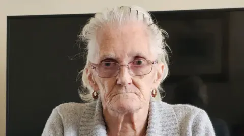 An elderly woman with shoulder length grey hair looks directly at the camera. She is wearing a grey jumper and glasses. She has an unhappy expression.