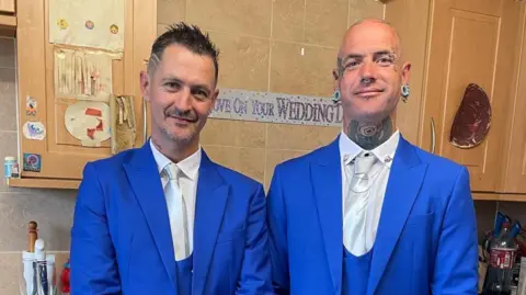 Family handout Both men are standing in a kitchen with pale pine cabinets wearing bright blue suits and white shits and tie. Behind them is a sign on the wall that appears to say "Love on your wedding day". Daniel Coleman is standing on the left of the image and is smiling with his head slightly inclined towards his brother. He has short dark hair with two lines shaved into the side. Tony Nye is also smiling and has facial piercings and parts of tattoos visible on his bald head and on his neck.
