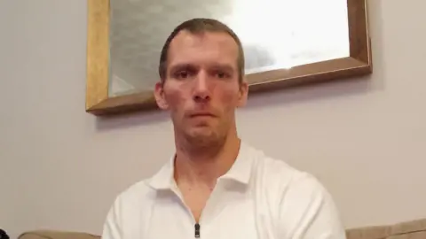 Leigh Walker has short dark hair and beard. He is sitting in a poorly lit room on a cream sofa. Behind him is a wood framed mirror. He is wearing a white polo shirt with a zip front. 