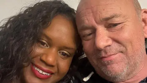 A selfie picture of Kacey a woman with black hair and Lee a man with short grey hair, smiling for the camera.