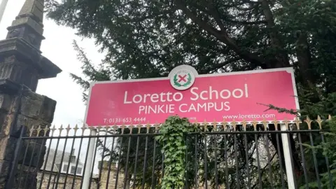 A sign outside Loretto College Pinkie Campus stands over an iron fence next to a stone gatepost.