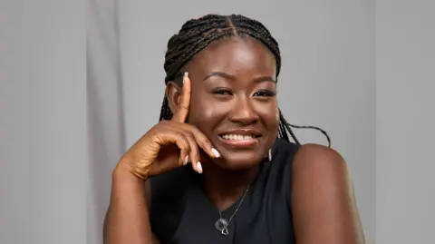 Jemima Antwi/ReCorn Imagery A woman with braids is smiling and leaning her face on her hand. She is wearing a black top and is photographed in front of a grey background. 