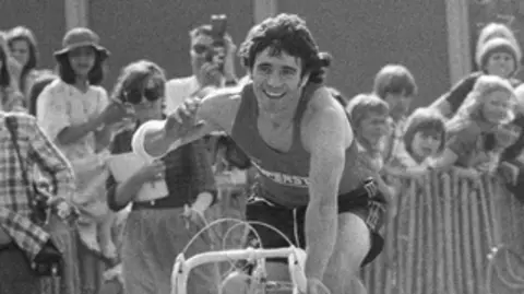 A black and white photo of Kevin Keegan riding a track bike and smiling in celebration of victory.