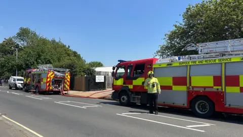 Alice Cunningham/BBC Two fire engines parked on the side of a road on a sunny day. There is a lone firefighter dressed in a bright green coat and helmet as well as black trousers. 