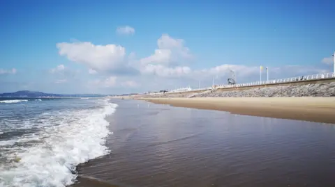 A sandy beach with the sea in front and a stone wall and while fence behind.