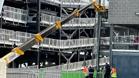 Helen Mulroy/BBC A group of people watch on as a large yellow telescopic handler is directed towards a gap in the car park where the vehicle fell from. A long metal barrier is hanging off the side of the building.