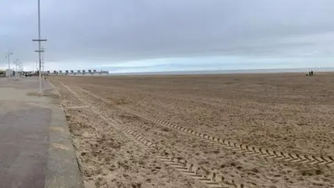 A view of the beach on a cloudy day.  A vehicle has left tracks in the sand in the foreground. Part of the promenade is in the left hand side of the picture.  The beach is deserted apart from a couple walking their dog in the distance.