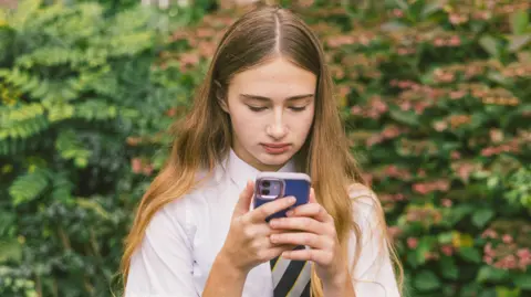 A girl in a white school shirt and striped tie with long brown hair holds a phone with a blue phone case. She stands looking at her device with a bush in the background. 