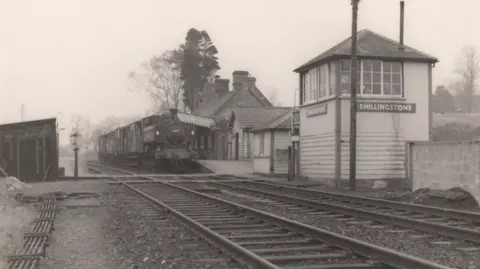 Paul Antell Collection An old black and white photo shows a steam train sitting in the station with the wooden signal box in the foreground.
