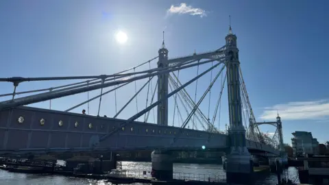 Albert bridge stretches over The River Thames. There is a blue sky and the sun is shining.