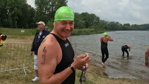 Nick Parkes is pictured in a wetsuit and green swimming cap with goggles in hand by the side of a lake where other swimmers are beginning to get in the water