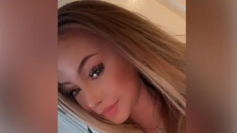 A slanted, selfie image of a young woman with long blonde hair. She is wearing a necklace and looking directly into the camera. 