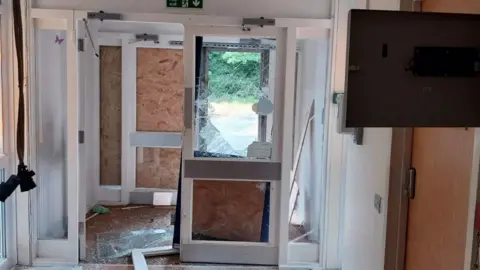 An abandoned building which is in need of repair. There are smashed glass windows and lots of boards up in place of where glass should be. Debris is on the floor.