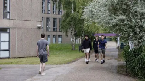 Four male students walk through York University campus. They are wearing shorts and T shirts. The apartment buildings are grey with tall windows.