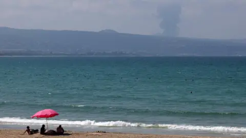 Reuters A family sits on the beach in Tyre, Lebanon, as smoke rises on the Lebanese side of the border with Israel during heavy gunfire between Hezbollah and the Israeli army on August 25, 2024