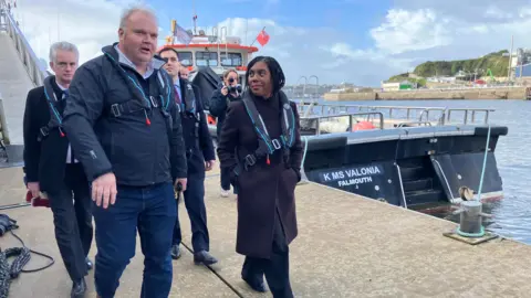 Kemi Badenoch walking to the right of a man. There are two other men behind her. She is walking on a dock alongside Plymouth's marina.