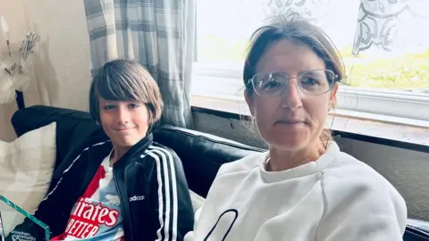 A boy with dark brown hair and navy and white sports jacket is sitting next to a woman. She is wearing a white sweater and is wearing glasses. 