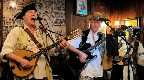 Three male musicians dressed as pirates perform at the Shakespeare pub in Bristol city centre. All three are playing guitars and wearing pirate hats. One of them is singing into a microphone