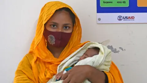 A woman is holding her child in her arms. She is wearing a face covering, and has an orange scarf draped over her head. The child has its eyes closed and is wrapped in a white cloth. 