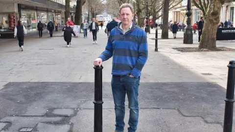 Andrew Booton standing in the middle of the promenade, wearing dark blue jeans and a blue striped jumper. He has short grey curly hair and is looking at the camera with a serious expression. His right hand is resting on top of a black bollard, while his other hand is in his jeans pocket. Behind him you can see shoppers walking down the road. Beneath his feet is a large patch of tarmac with has been laid to replace the broken paving stones with cover the rest of the street, 