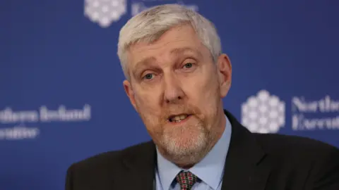 PA Media John O'Dowd speaking. He's wearing a blue shirt, red and navy patterned tie and a black blazer. He has grey hair and a beard.