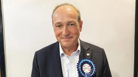 A man wearing a suit and a blue rosette. He is looking towards the camera.