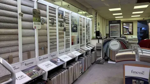 The inside of the carpet shop. There are vertical stacks of carpet samples on a display on the left hand side. It also has samples hanging underneath them. To the right, there are booklets of carpet samples that you can flip through.