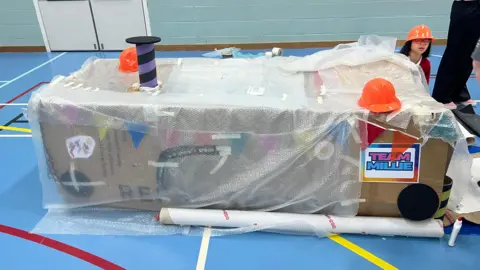 A large cardboard box which has been decorated with colourful bunting and drawings by children, and wrapped in bubble wrap. It is on the blue floor of an indoor sports centre.