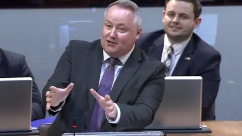 Darren Millar in the Senedd chamber criticising Eluned Morgan for not giving him her definition of a woman. He is smiling and gesturing with his hands and has a Conservative colleague behind him who is also smiling