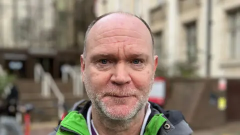 Simon Dedman/BBC A man - Andy Abbott - looking straight at the camera. He is wearing a green and grey coat and is standing in front of a set of steps outside a building.