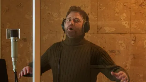 Gerard Butler stands in a sound booth wearing a khaki turtle neck. His mouth is slightly open looking like he is singing and his hands raised up. 