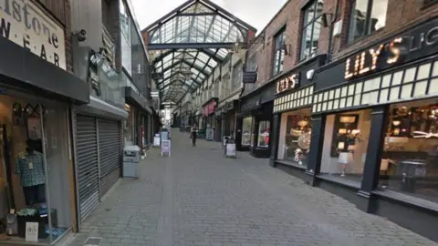 A cobbled street with shops lining each side. There is a glass roof covering some of the buildings. 