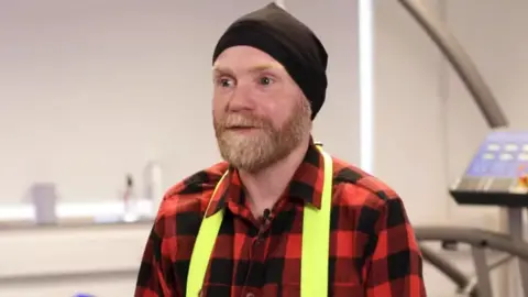 University of Worcester The man from the above picture with a slightly less bushy beard, wearing a black beanie hat and a red and black lumberjack shirt.