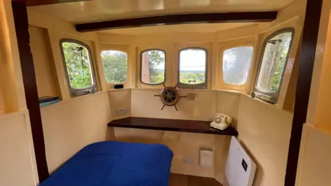 Ellen Knight/BBC The interior of the shed - which has cream painted walls and a wooden floor. There are six small windows at the front, and a small wooden ship's wheel beneath them. A single bed is in the bottom left corner of the image, with a bright blue duvet. 