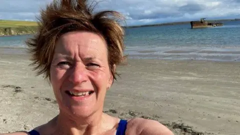 Linda Sinclair Selfie head shot of a woman on a beach with the sea and a ship wreck in the background