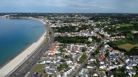 An aerial view of Victoria Avenue - a view of homes and streets along Jersey’s coastline, with a curved beach and coastal road beside the sea.




