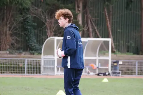 Lukas Colese A young man in a blue tracksuit on a training ground. There are yellow cones on the grass, and a glass dugout shelter in the background.